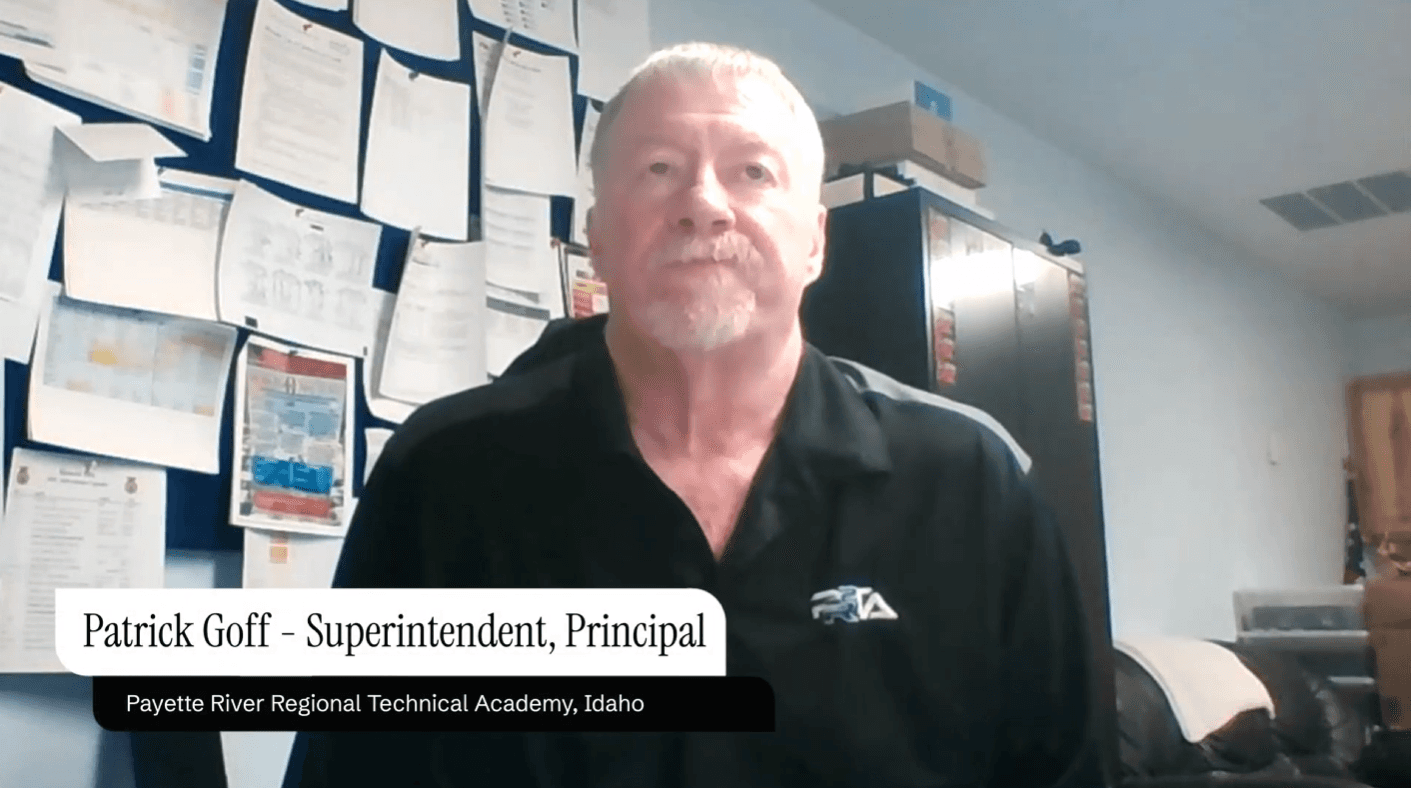 A man with short hair and a trimmed beard appears on camera, facing forward as if in a video call or recording. He is wearing a light blue collared shirt. The background shows a plain, light-colored brick wall, giving the setting a simple indoor environment such as an office or classroom.