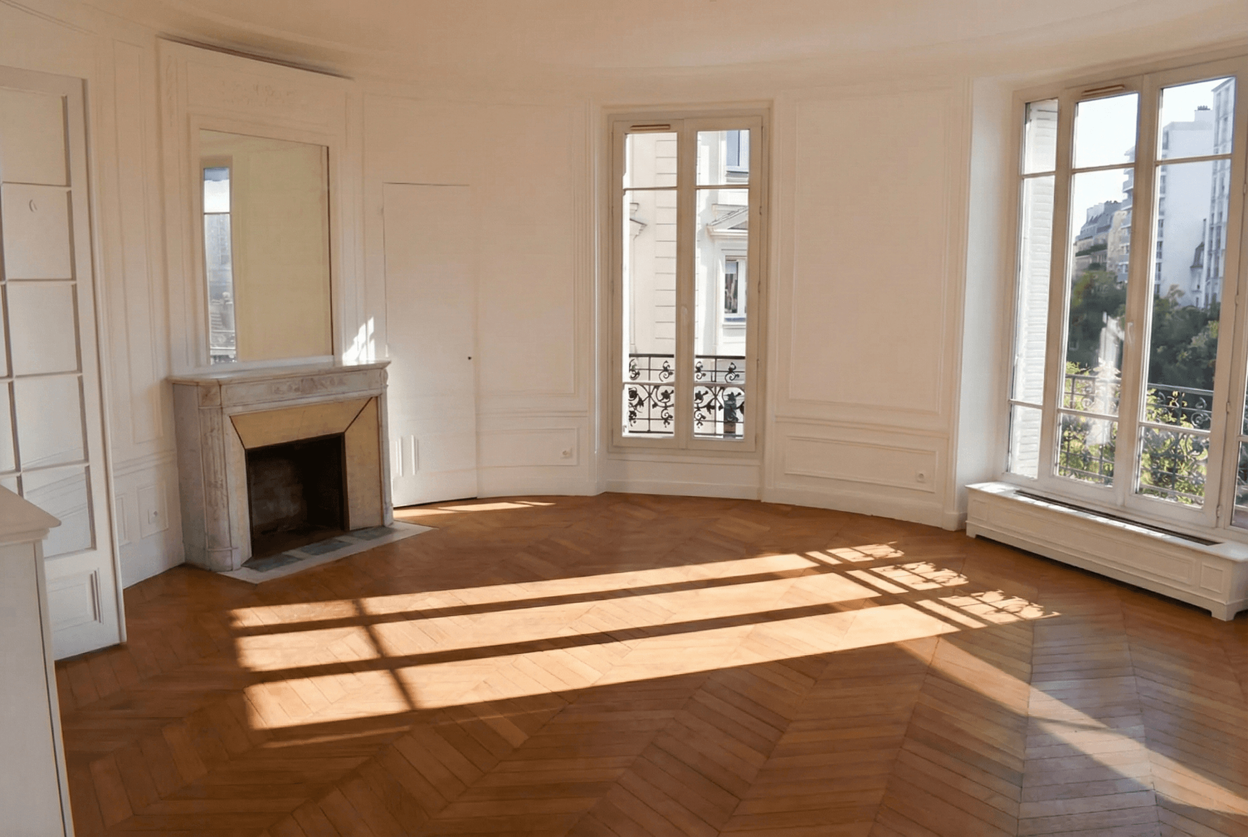 Grand salon haussmannien vide avec parquet point de Hongrie, cheminée ancienne en marbre et grandes fenêtres lumineuses.