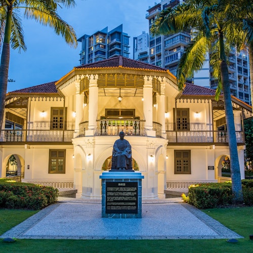 An illuminated two-story colonial building with a red roof, palm trees, and a statue in front, set against a backdrop of modern high-rises.