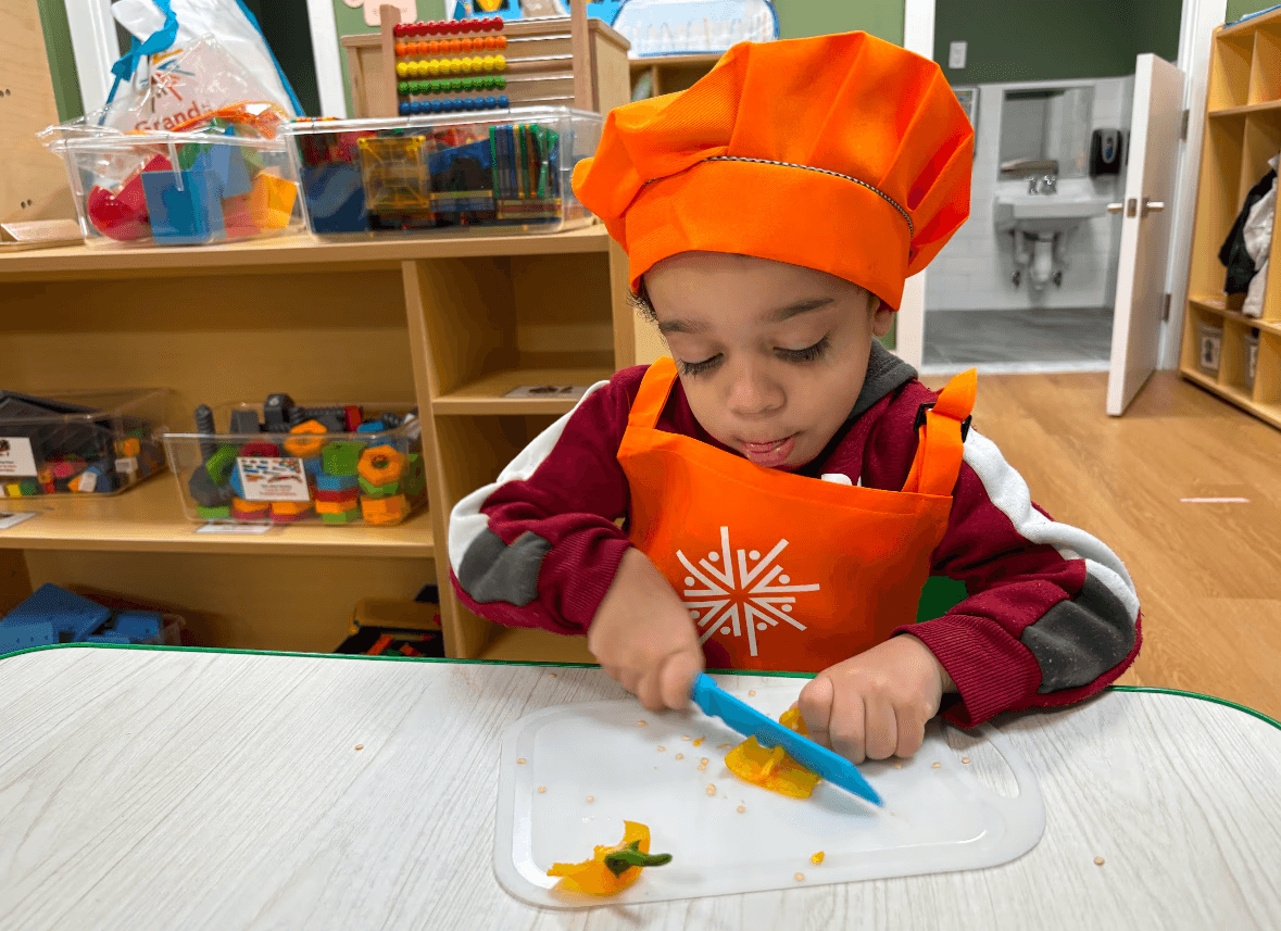 Preschooler in chef's clothes carefully uses a child-safe impelment to chop a pepper.
