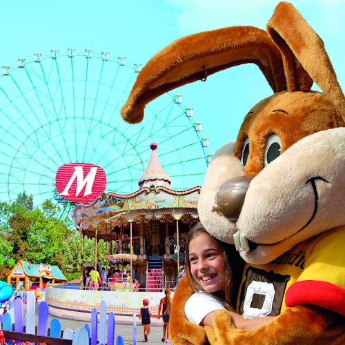 A smiling child hugs a large bunny mascot at an amusement park, with a Ferris wheel and carousel in the background.