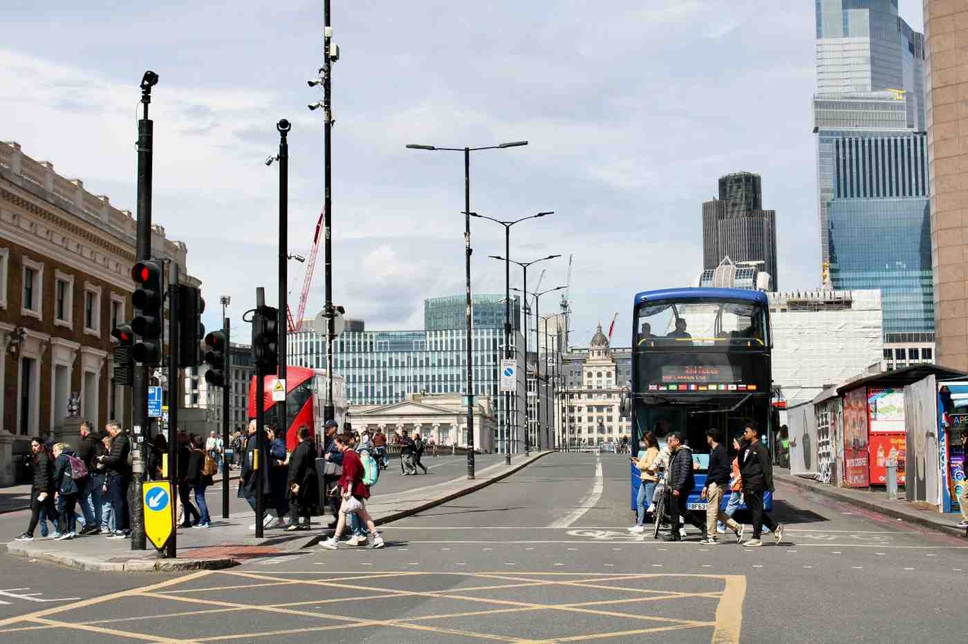 Pedestrians crossing a busy junction in central London with traffic lights, buses, and modern office buildings in the background.