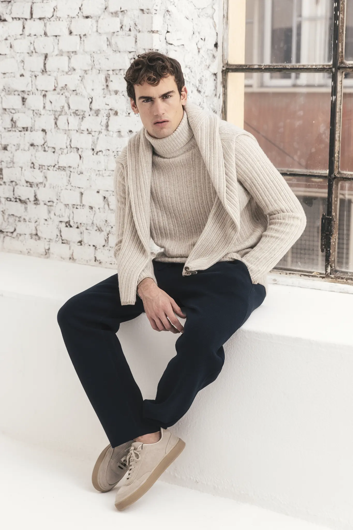 Man in ivory ribbed cashmere turtleneck and navy trousers, with matching cardigan draped over shoulders – Aida Barni FW25/26 collection.
