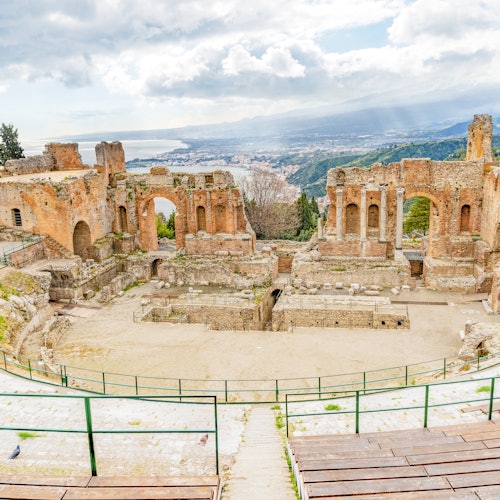 Ruines d'un ancien amphithéâtre en pierre avec des sièges en gradins et une vue pittoresque sur la côte et les montagnes en arrière-plan.