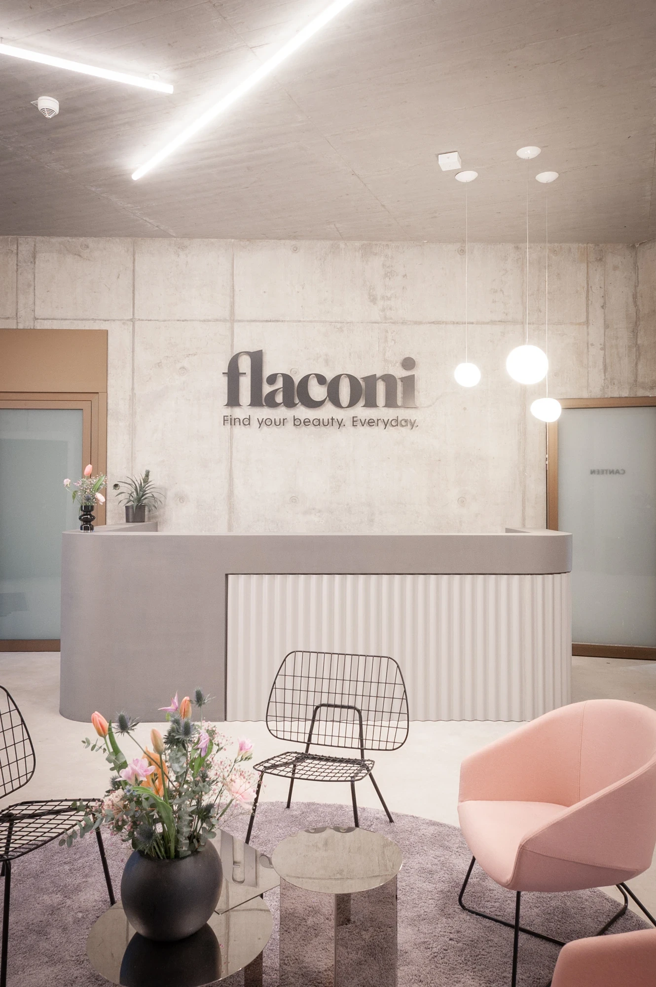 Flaconi reception area with the brand logo and tagline „Find your beauty. Everyday." on a raw concrete wall, ribbed reception desk and pastel pink lounge chair.
