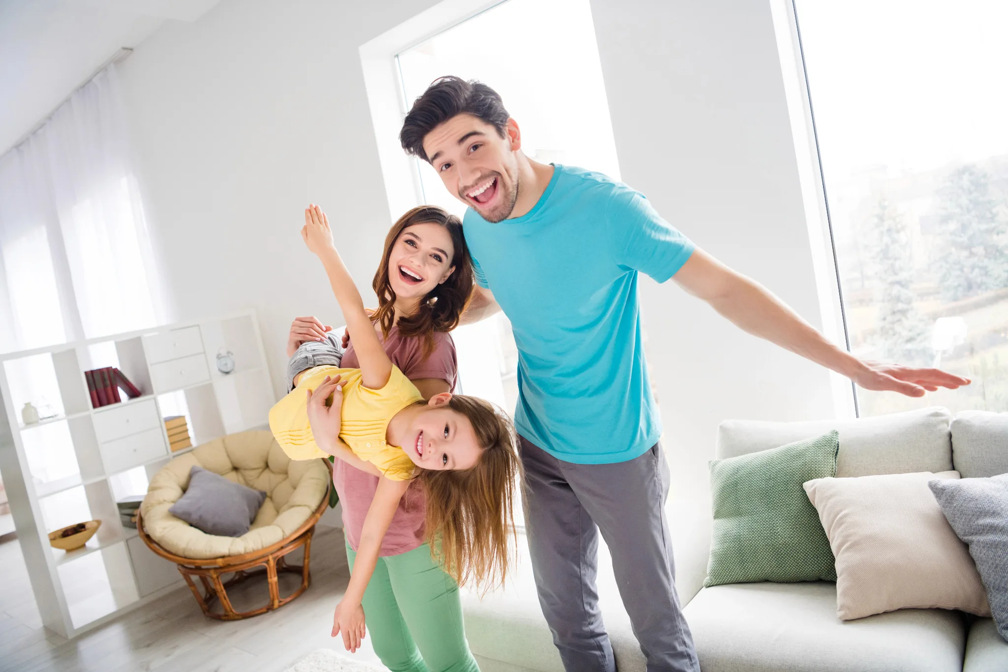 Happy family standing in a house. large windows, light from the outside reflecting inside the house. Mother holding a daughter with two hands up in the air like an airplane, husband's right arm on wife's shoulder.