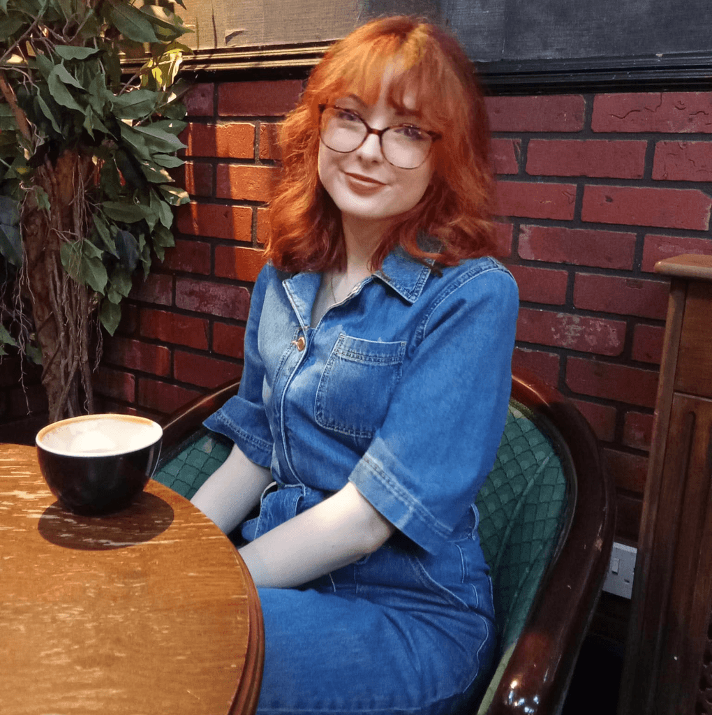 Amy sitting on a green chair with her back to a red bring wall, she is in a coffee shop and has a cappuccino on a wooden table in front of her.