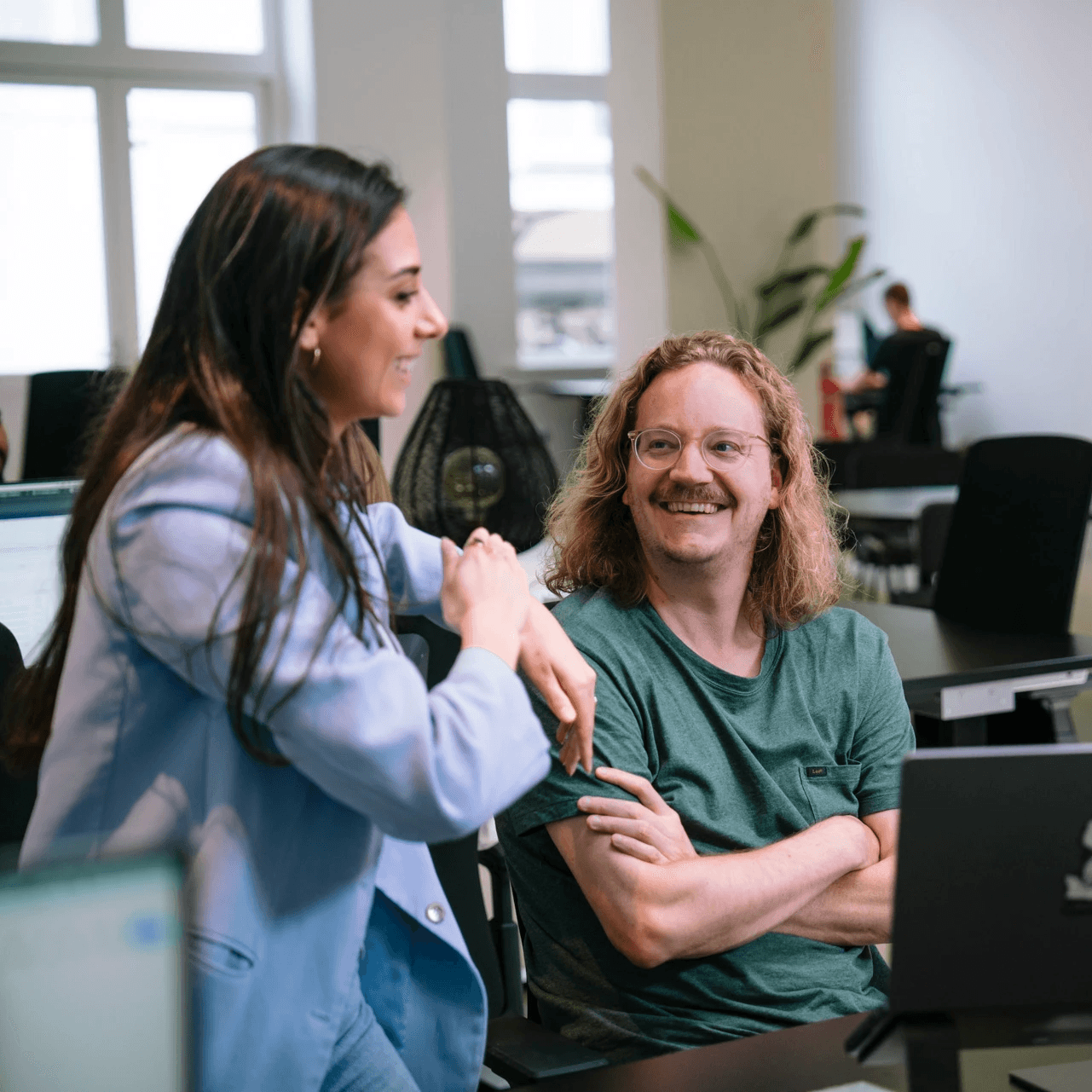 A woman in a blue blazer talks enthusiastically to a man with curly hair, both smiling in a modern office setting.