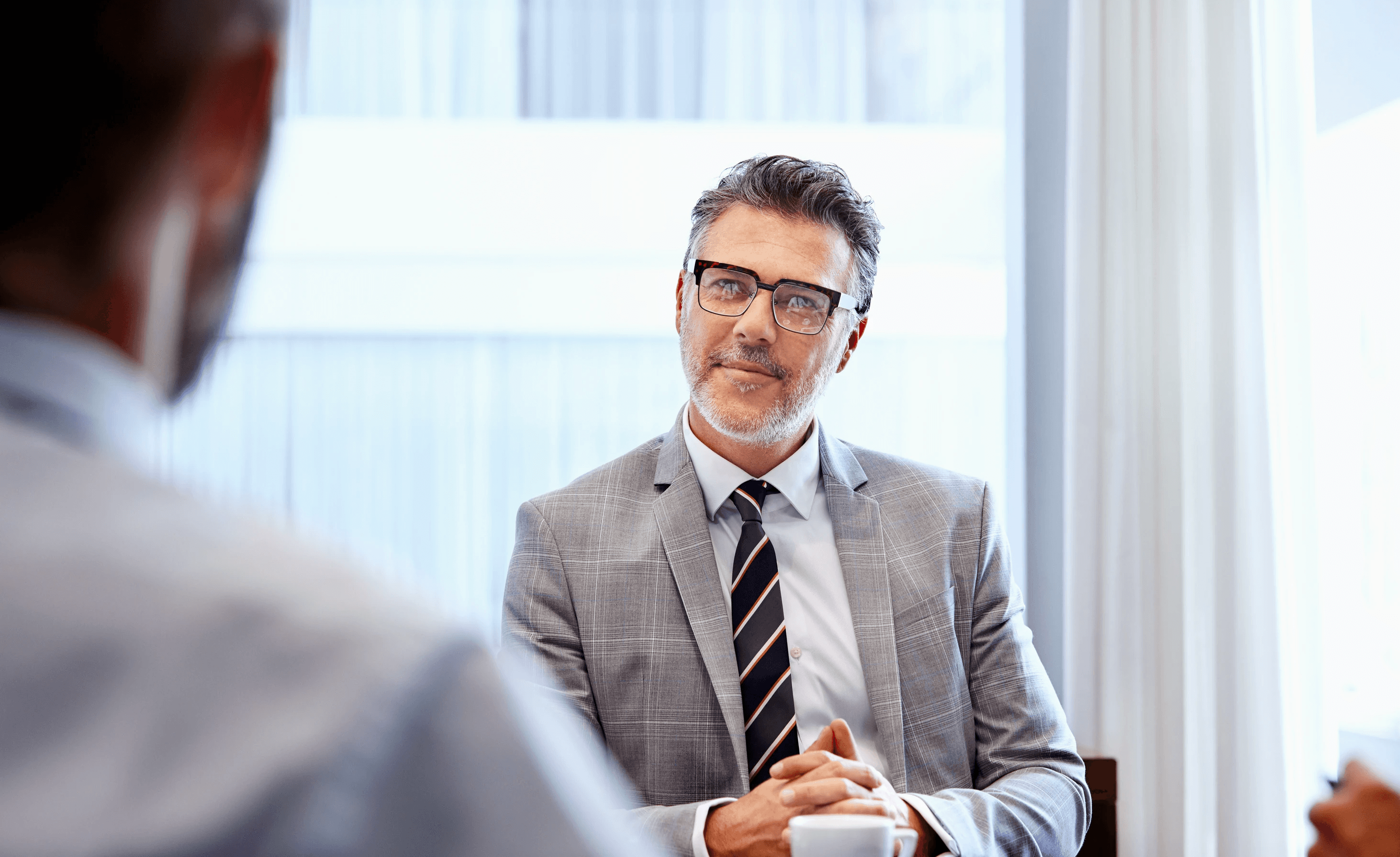 A retirement plan advisor in a gray suit listens during a client meeting, hands clasped at a conference table in a bright office