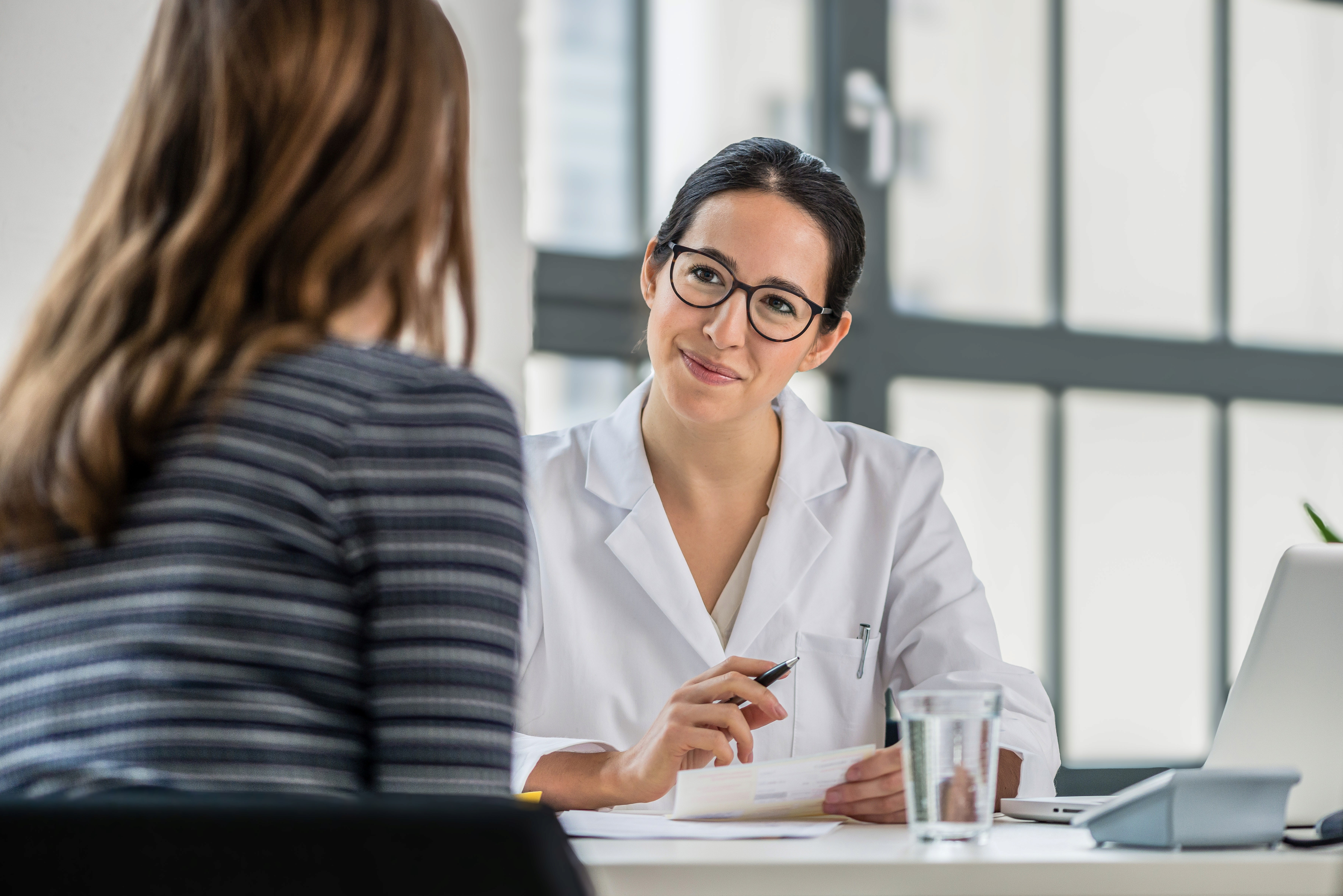 Man wearing an olive green jacket and black shirt, sitting and smiling in front of a light wooden background.