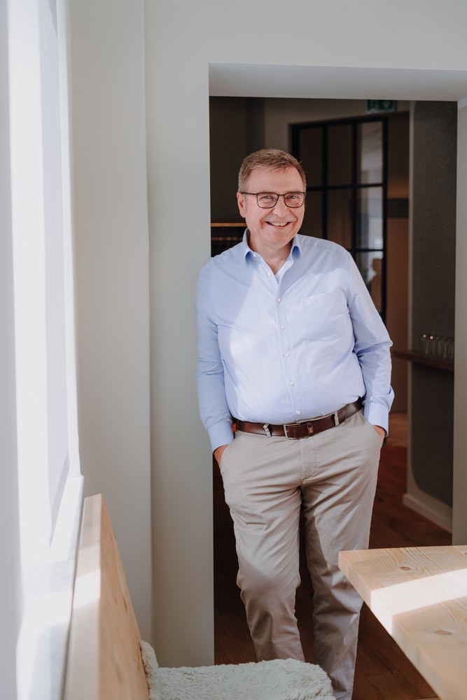 Männlicher Geschäftsführerin im hellen Hemd steht locker vor einem Fenster im Büro von SLB.