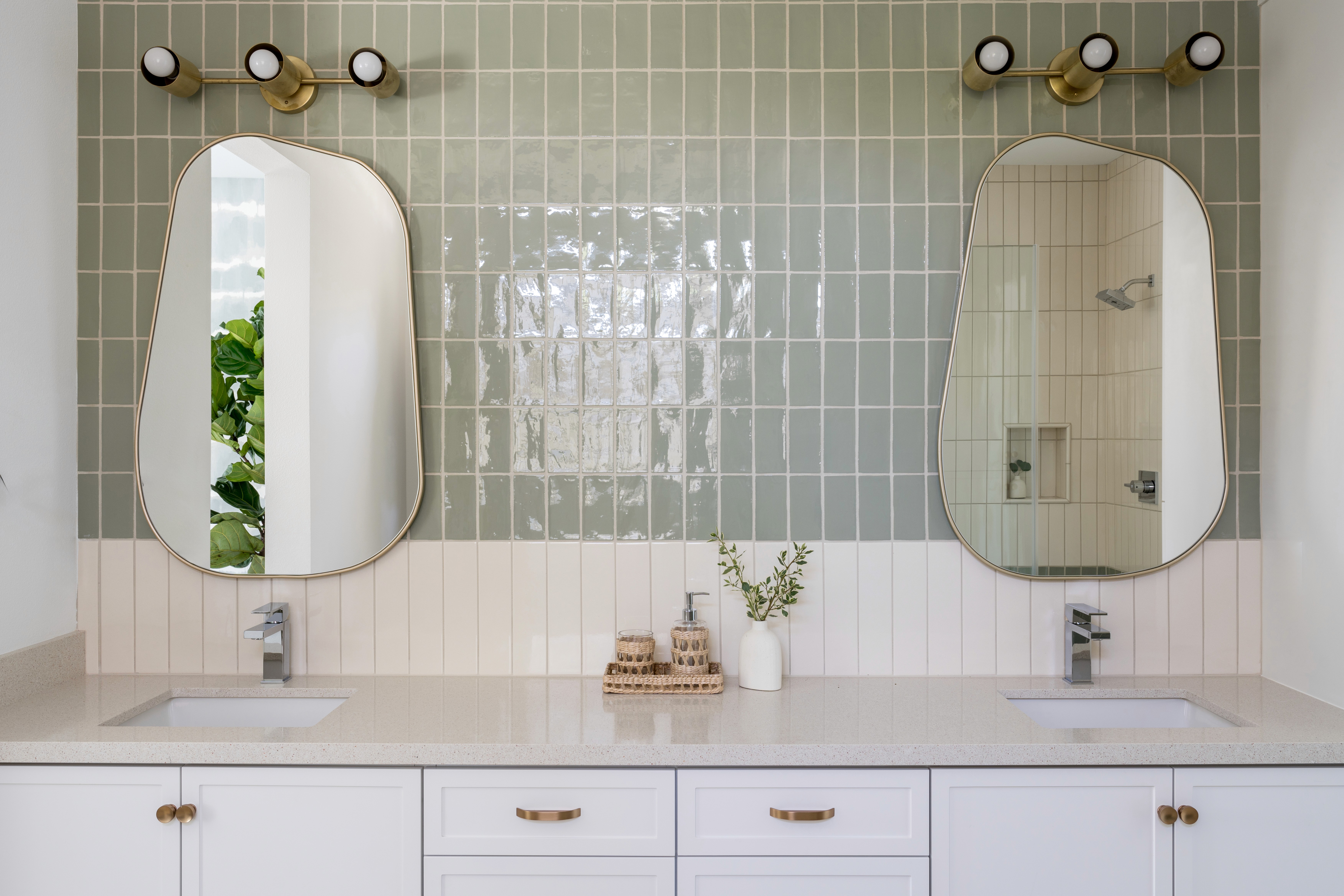Bathroom with dual vanity sinks, gold framed rounded vanity mirrors, sage green tiled wall, white cabinetry with brass finishes, and three-bulb, brass vanity lighting  by Jubilee Interiors in Arcadia, California