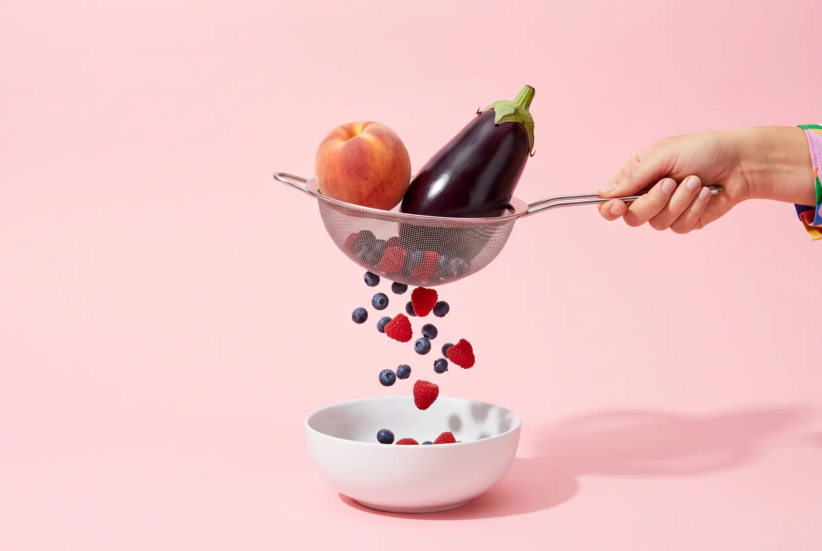 Kitchen sieve letting blueberries fall through while a peach and an eggplant stay caught on top, a metaphor for a DNS filter that blocks Reddit NSFW