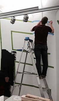 A photo of a man standing on a leader and sticking a blue green duct tape so that letters can be formed and painted between the line.
