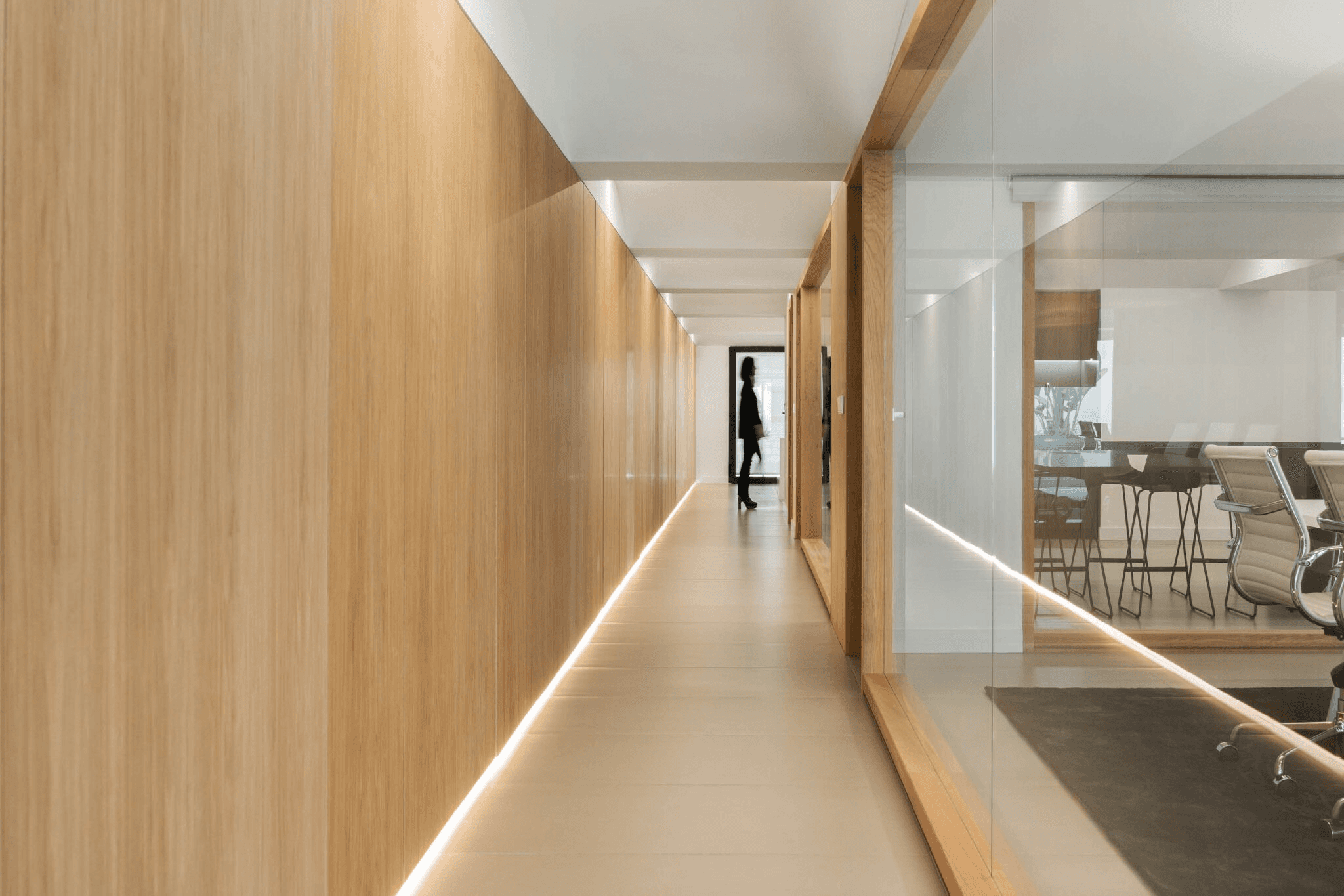 Modern office hallway with wooden paneling and glass walls, leading to a conference room with sleek office chairs, featuring a minimalistic and contemporary design.
