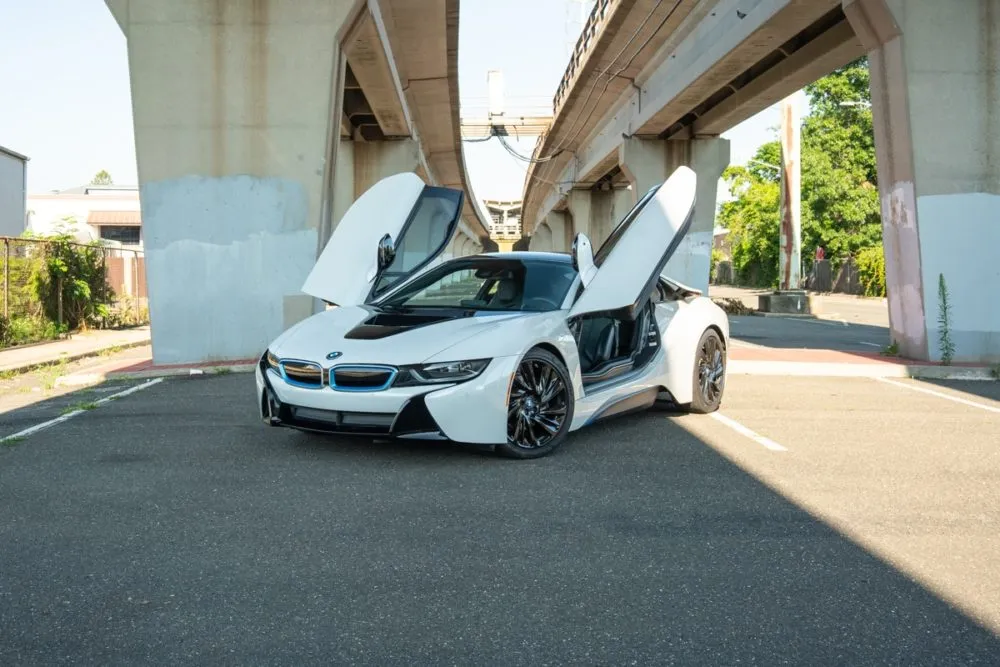 Front angle view of a white BMW i8 parked beneath an overpass in Miami, dihedral doors open for dramatic effect.