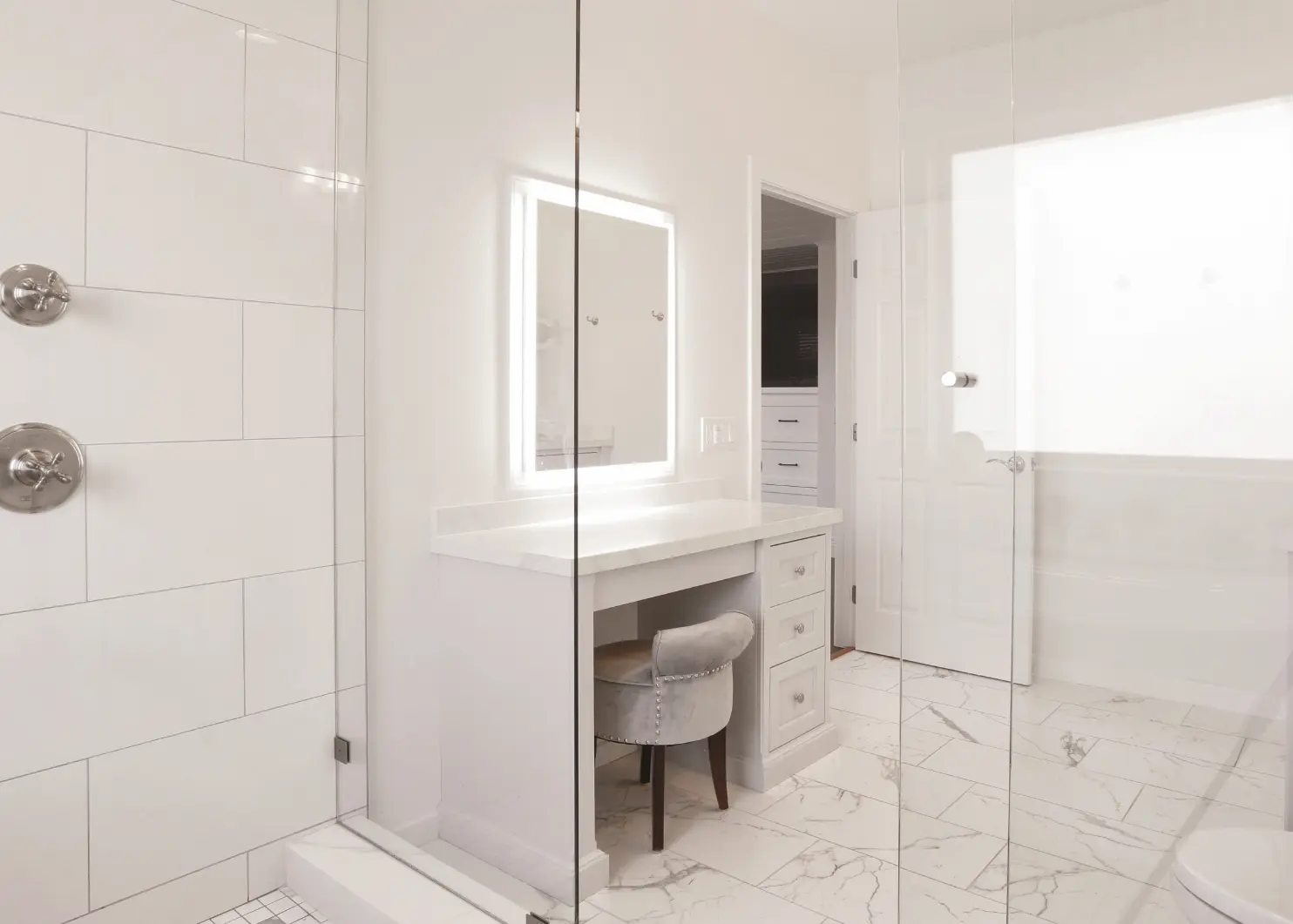 View of makeup vanity area with led mirror and velvet chair seen from the walk-in shower.Primary bathroom with muted tones, double vanity, makeup vanity, and spacious walk-in shower and marble tile. Photo by Chris Darnall.