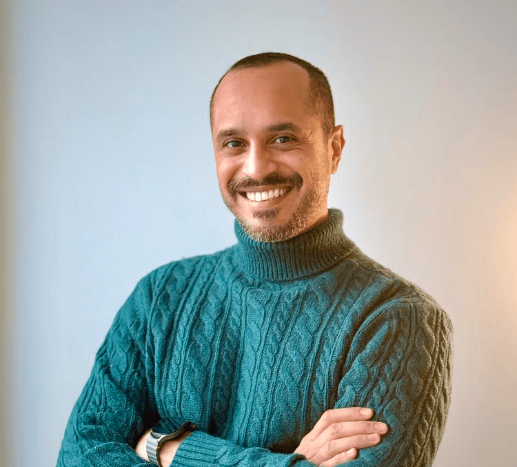 A serious-looking person with medium-length wavy hair, wearing a black coat over a brown sweater and tie, exudes a classic, sophisticated style.