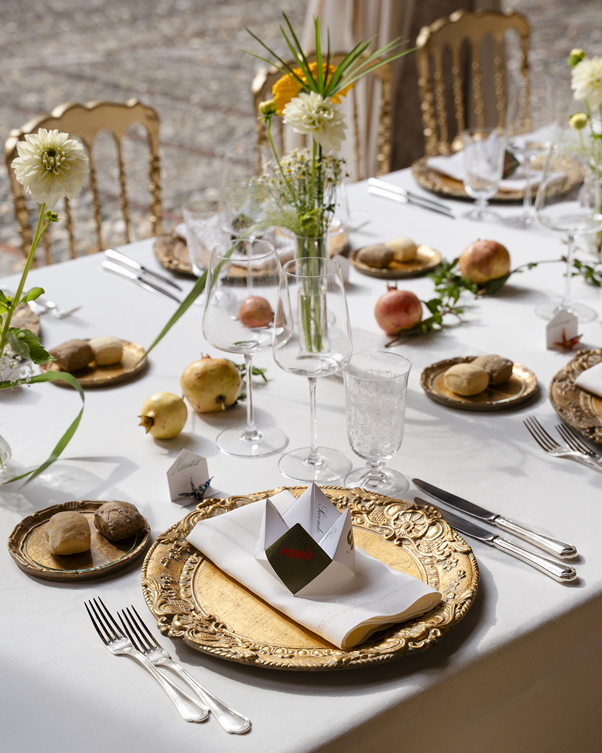 Dettaglio della mise en place con bicchieri, fiori in vasetto, frutta e pane su tovaglia bianca