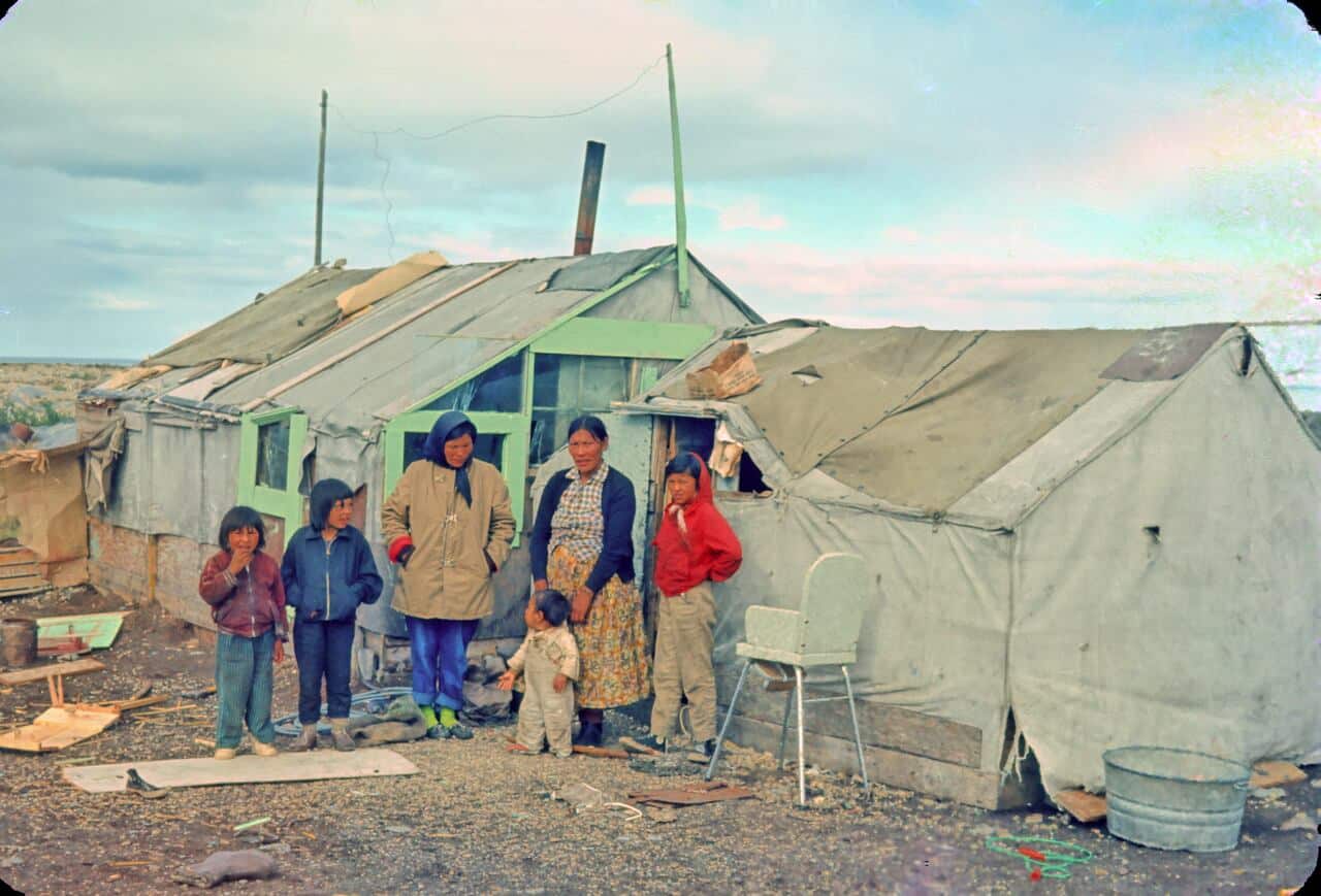 An archival colour photograph: a family stands outside a canvas tent and a small wooden shelter.