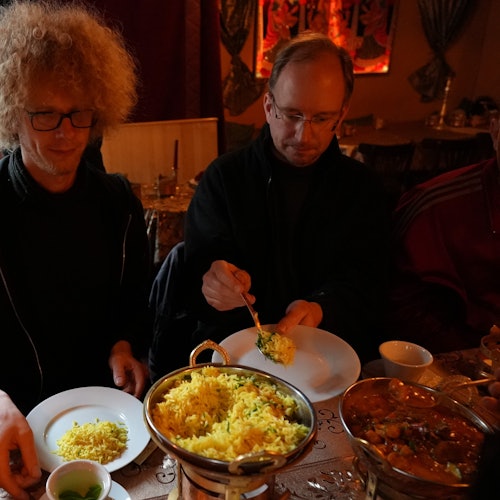 Two men are seated at a table, serving themselves from communal dishes of yellow rice and curry.