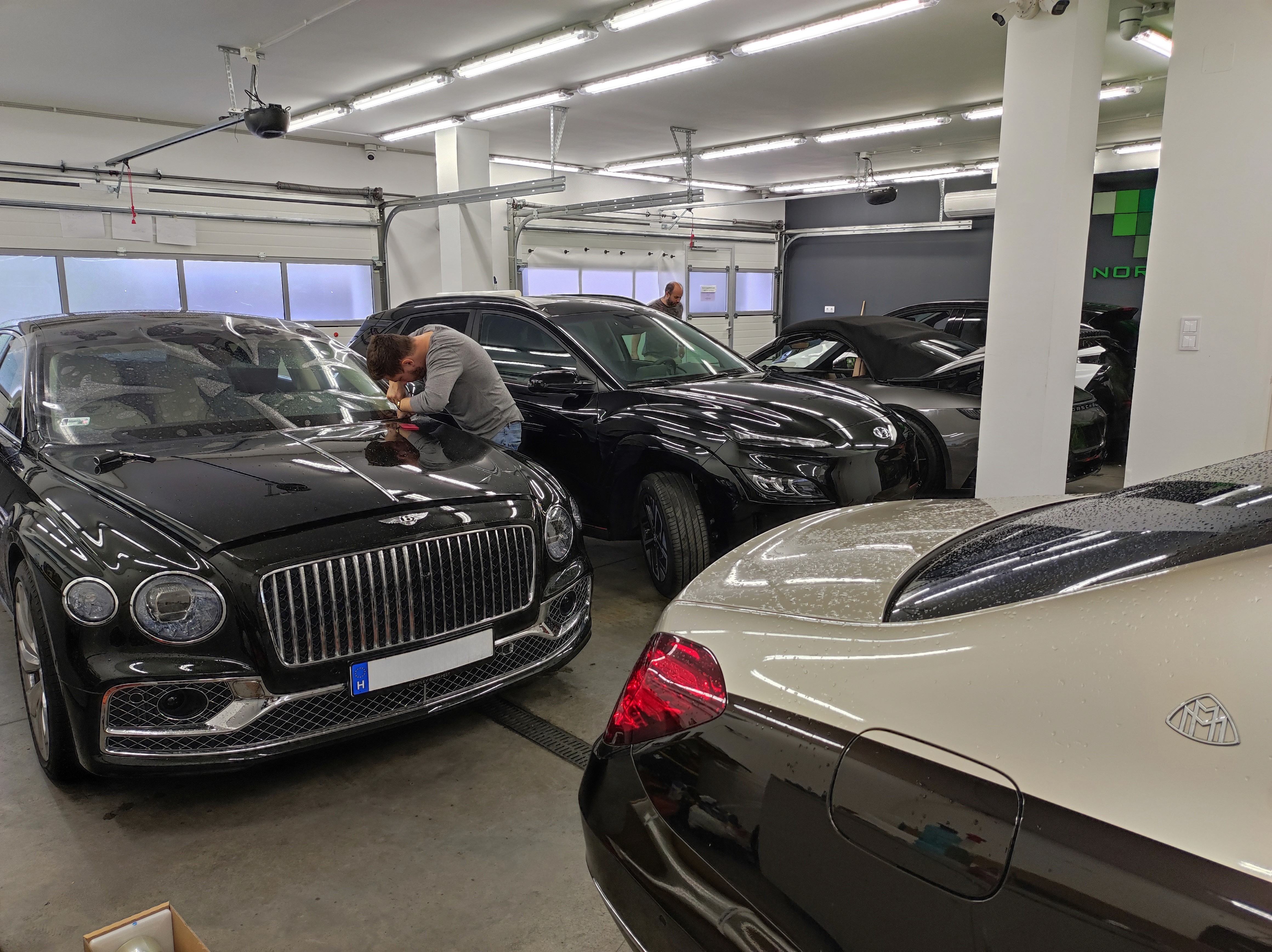 Karosszériavédő fólia (PPF) precíz applikálása egy fekete Bentley motorháztetején, az előtérben egy kéttónusú Maybach S600 luxusautóval.