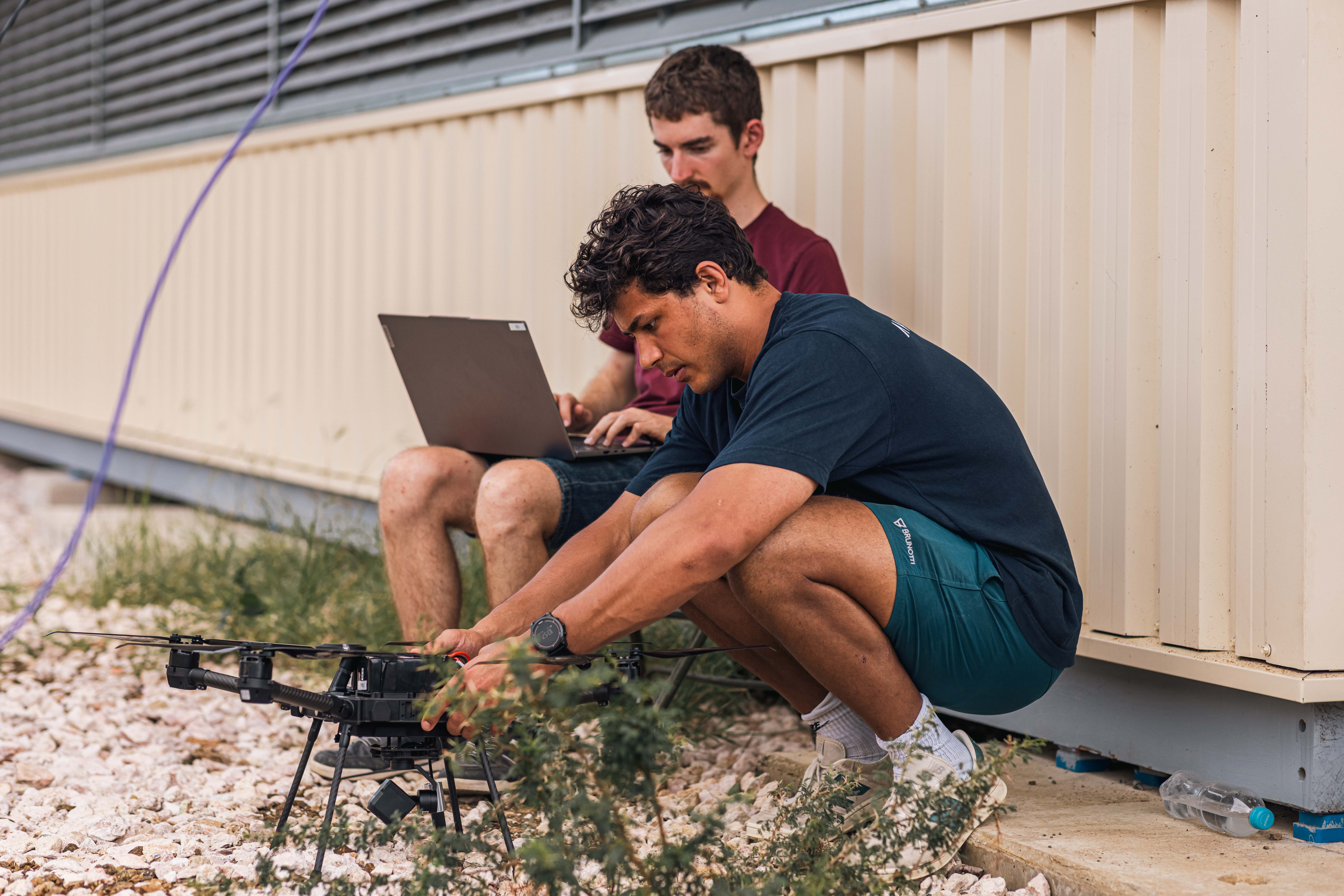 Two men are sitting on the ground near a beige building, one operating a laptop and the other adjusting a black drone, surrounded by gravel and sparse vegetation.
