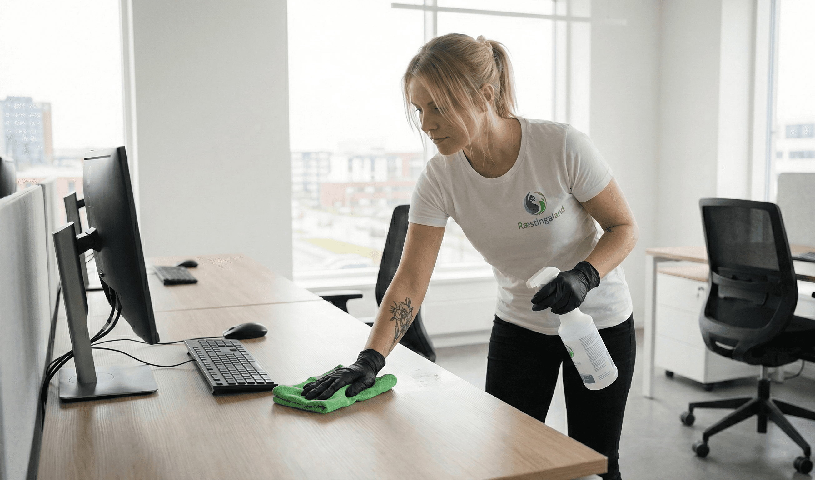 Starfsmaður Ræstingaland að þurrka af skrifborði á bjartri skrifstofu.