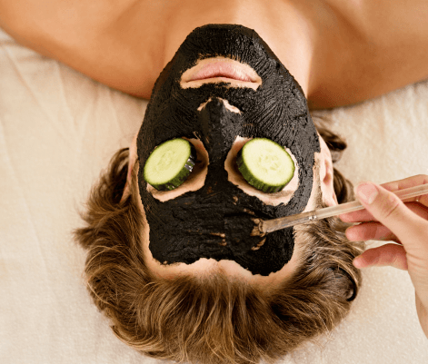 A person is lying down with a black facial mask applied to their face, and cucumber slices placed over their eyes. A hand is seen applying more of the mask with a brush. The setting appears to be a spa or skincare treatment area, with a neutral background and soft lighting.