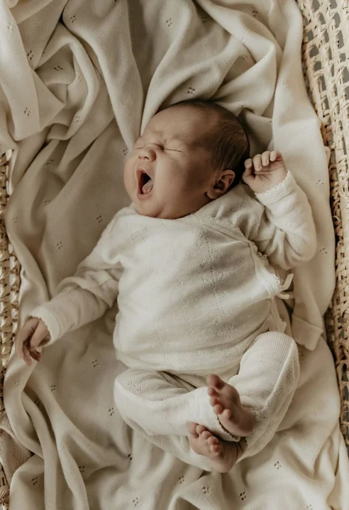 Yawning baby in a white outfit on a beige blanket