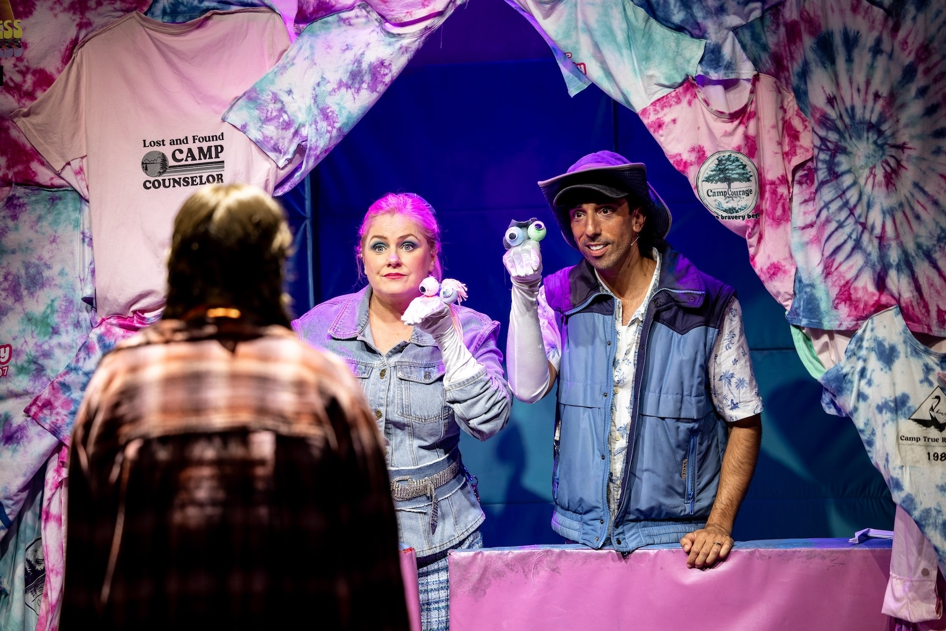 Two performers stand behind a makeshift puppet booth decorated with tie-dyed T-shirts, each holding a hand puppet with googly eyes. A third performer, seen from behind, faces them as they perform.
