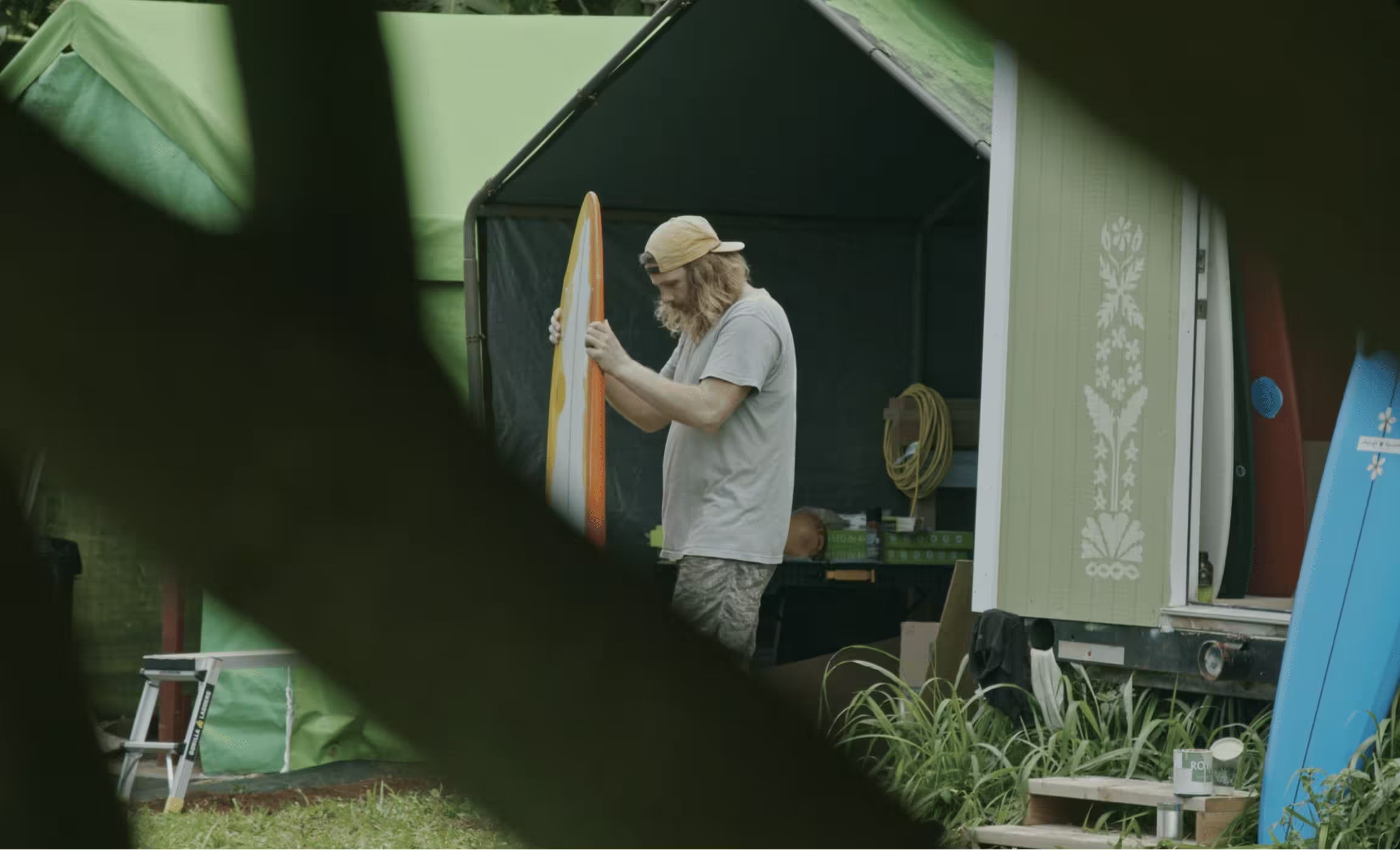Man holding surfboard outside green beach shack.