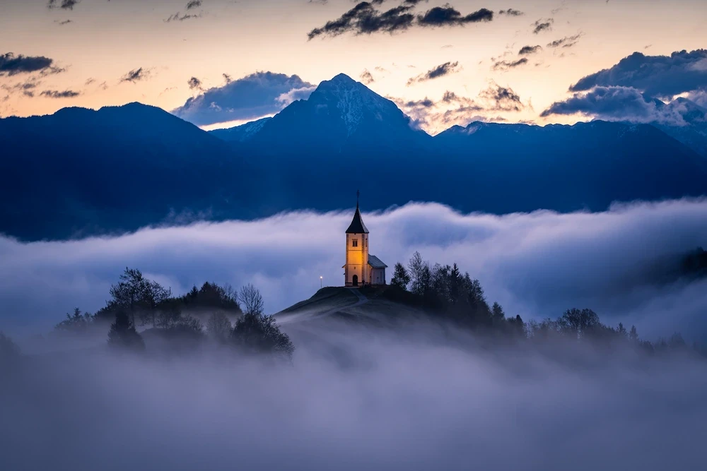 The iconic hilltop church of Jamnik in Slovenia, appearing as an island above a dense layer of low-lying fog during blue hour..