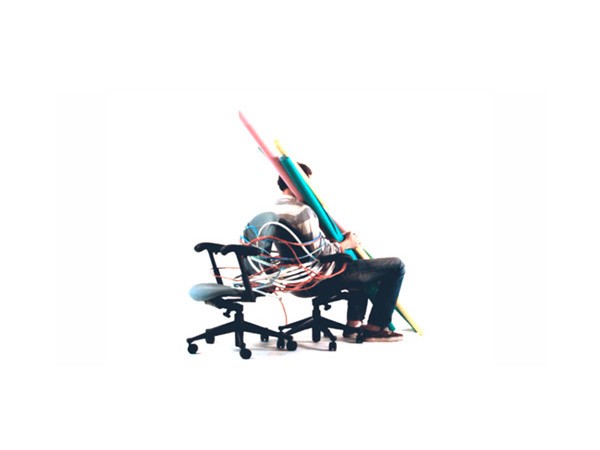 A person sits in a black rolling office chair with colorful cables draped over their head and shoulders. The chair has five wheels and is positioned against a plain white background. The person is wearing a light-colored shirt and dark pants, and their head is turned slightly to the right. The cables are multicolored and appear to be connected to various devices or equipment, though specific devices are not visible in the image.