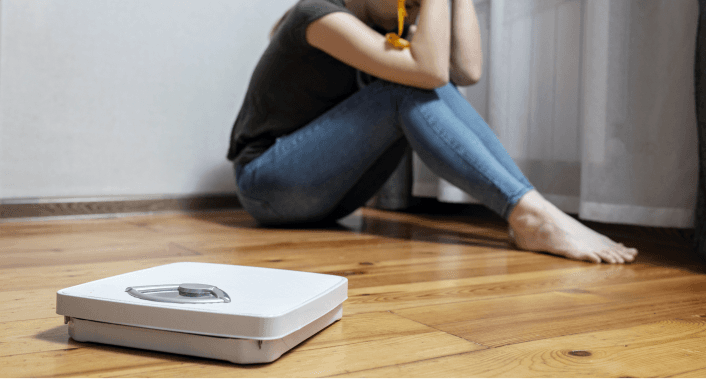 Person sitting on floor near scale, appearing distressed about weight.