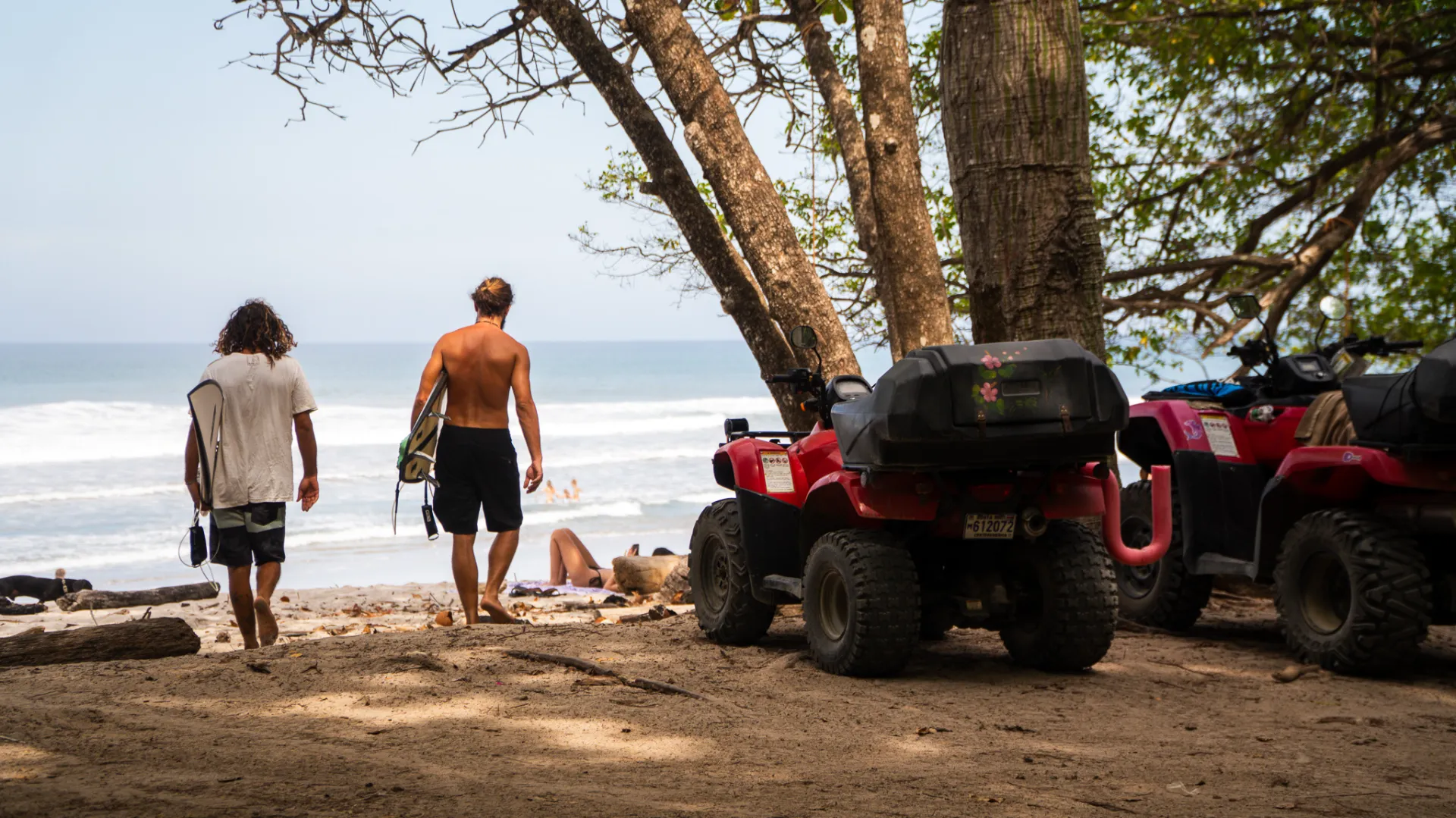 Surfers walking to Playa La Lora in Santa Teresa past ATVs parked under the trees
