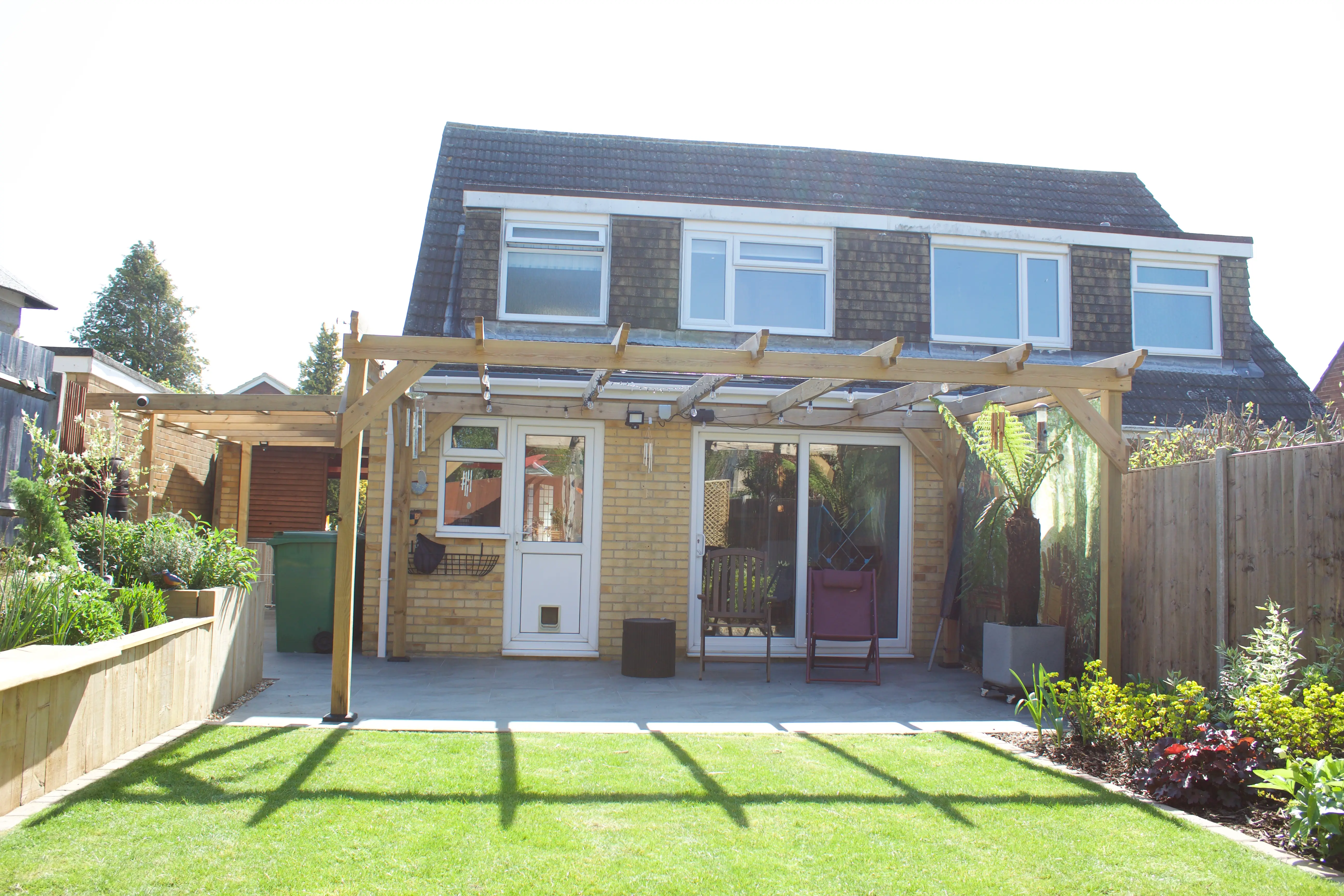 A modern house with large windows, a patio area, and a well-maintained garden in bright sunlight.
