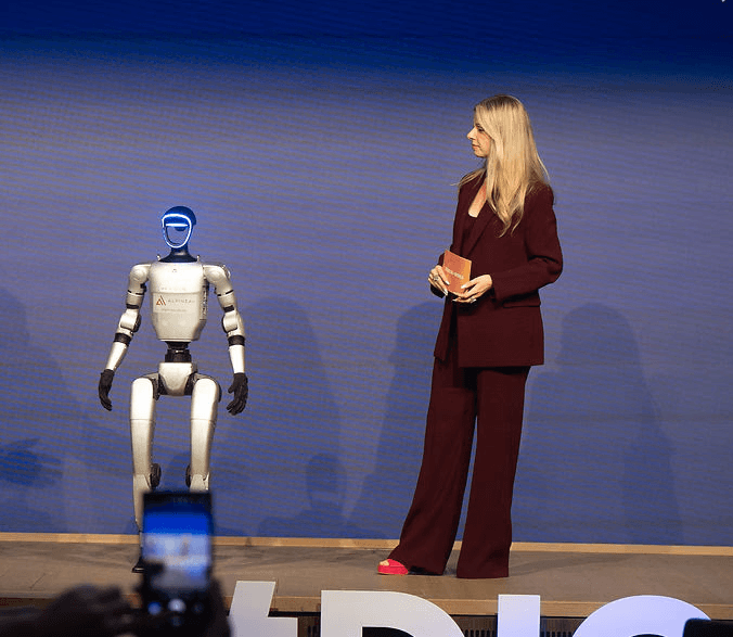 Moderatorin Désirée Lehmann im dunkelroten Hosenanzug auf einer Bühne mit einem Roboter.