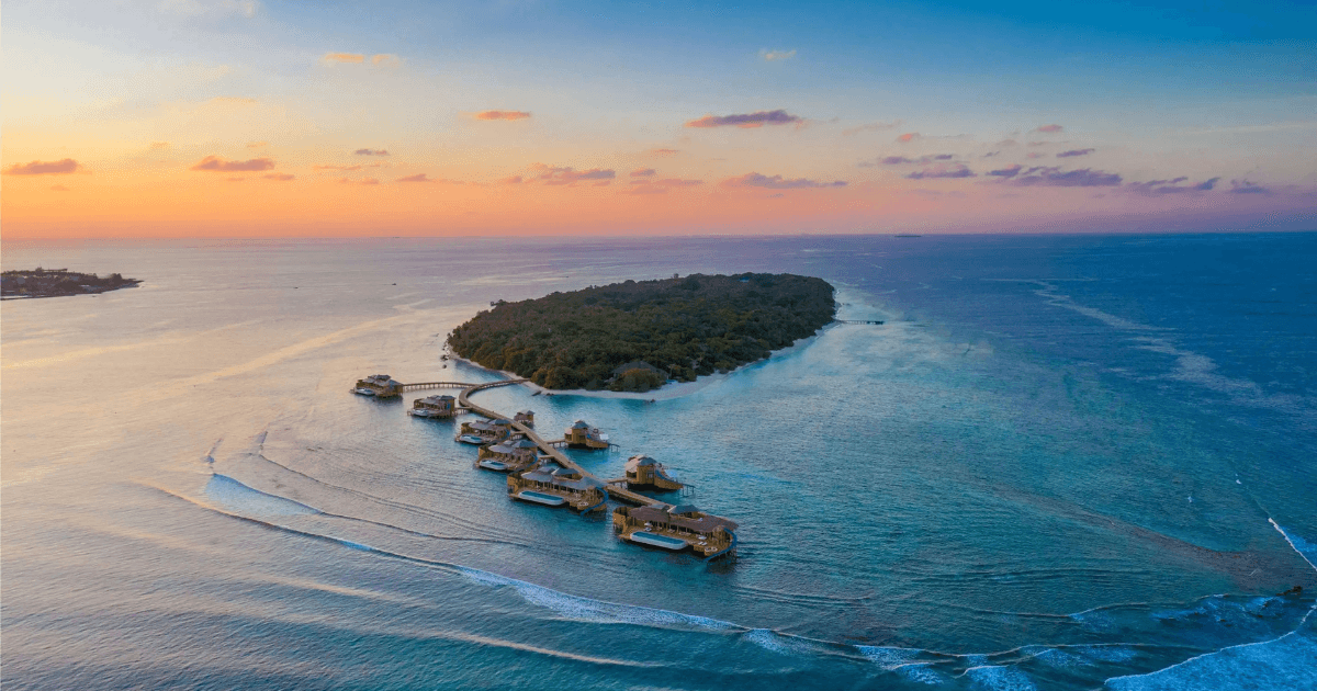 Aerial view of an island resort at sunset with villa houses floating above water extending into the ocean.