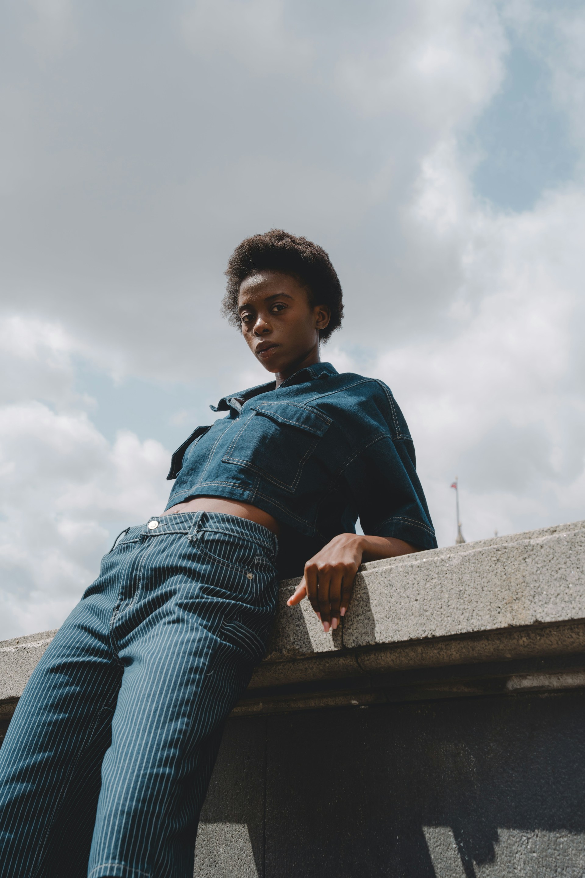 A close-up, low-angle shot of the woman leaning back against a stone wall under a cloudy sky. She is looking directly at the camera with a neutral expression.