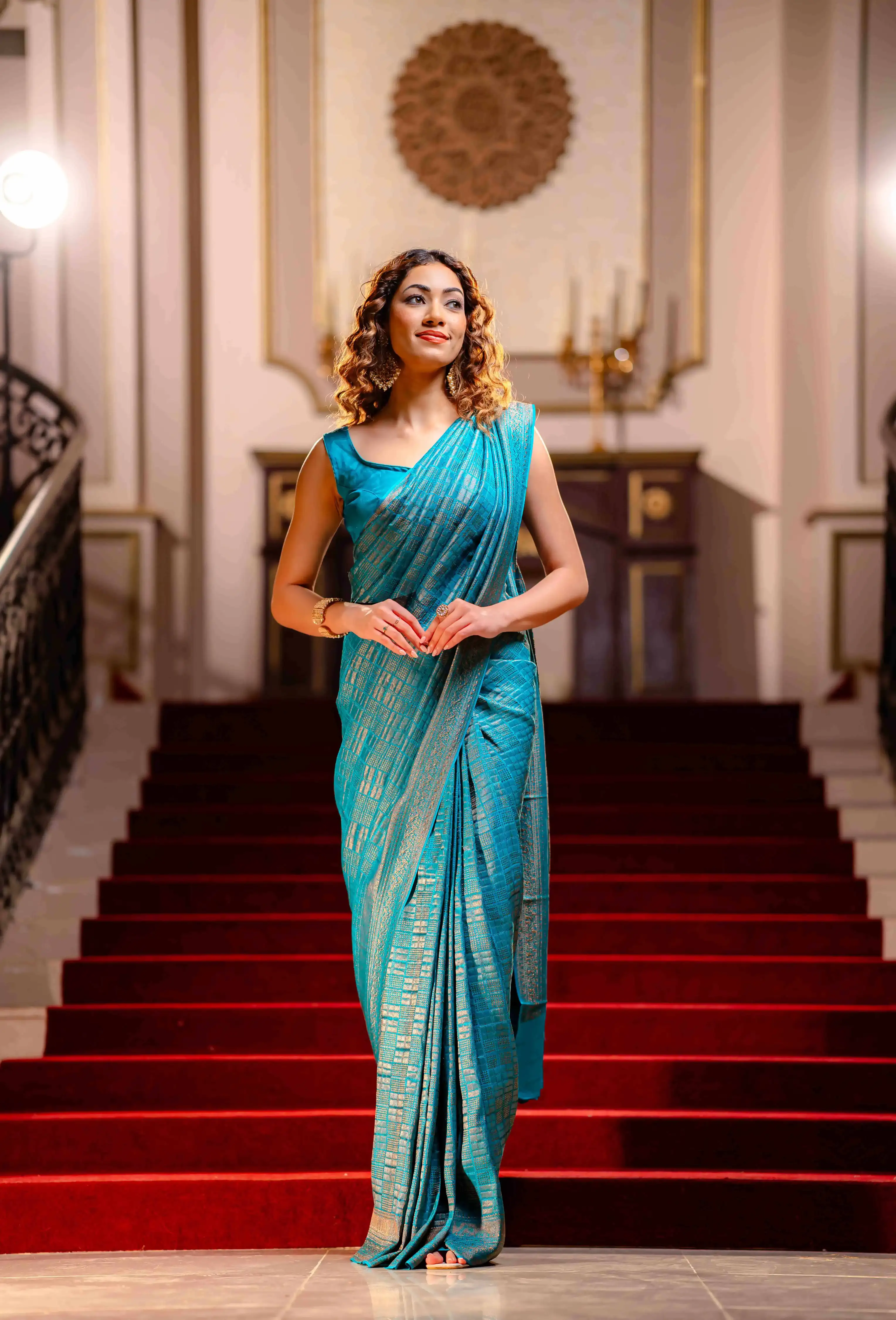 A woman wearing a blue saree from That Saree Shop, standing gracefully on a stair in an elegant room, highlighting the saree’s luxurious weave and classic festive charm.