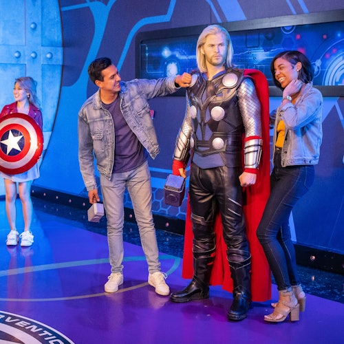 两个人在室内与雷神雕像合影，而背景中的一位女孩手持美国队长的盾牌。