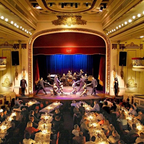An elegant theater scene with couples dancing onstage. Audience members dine at candle-lit tables, watching the performance.