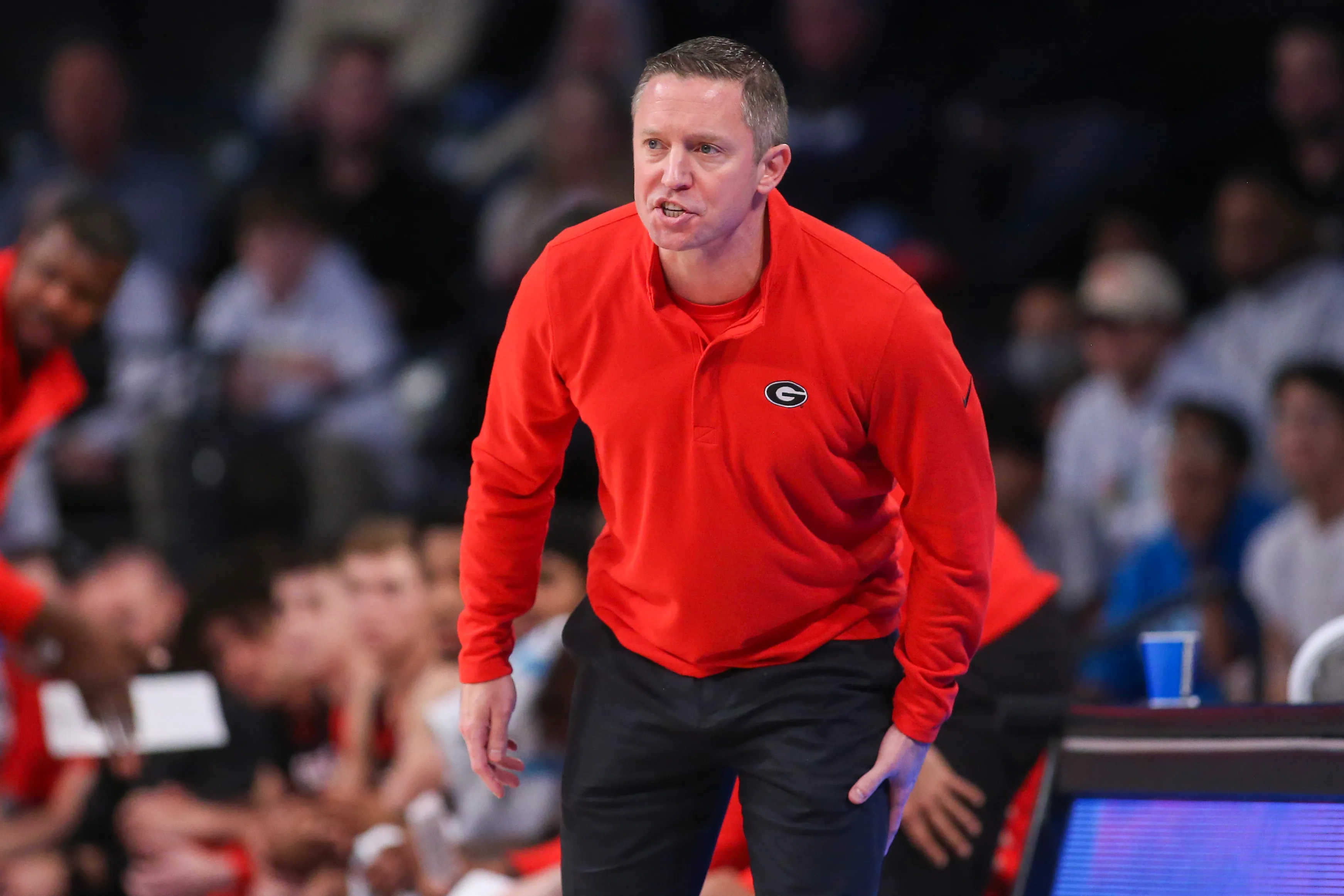 A basketball coach wearing a red Georgia Bulldogs pullover leans forward and shouts instructions during a game, with players and spectators visible in the background.