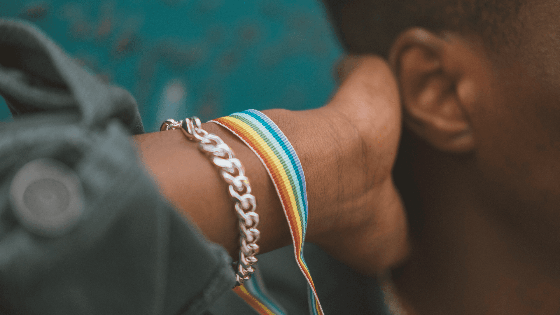 Close-up of a hand reaching towards an ear, adorned with colorful bracelets against a teal background.