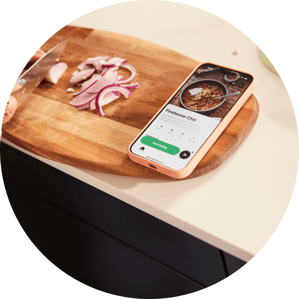 Smartphone on a wooden cutting board displays a "Firehouse Chili" recipe while sliced red onions and a knife sit nearby