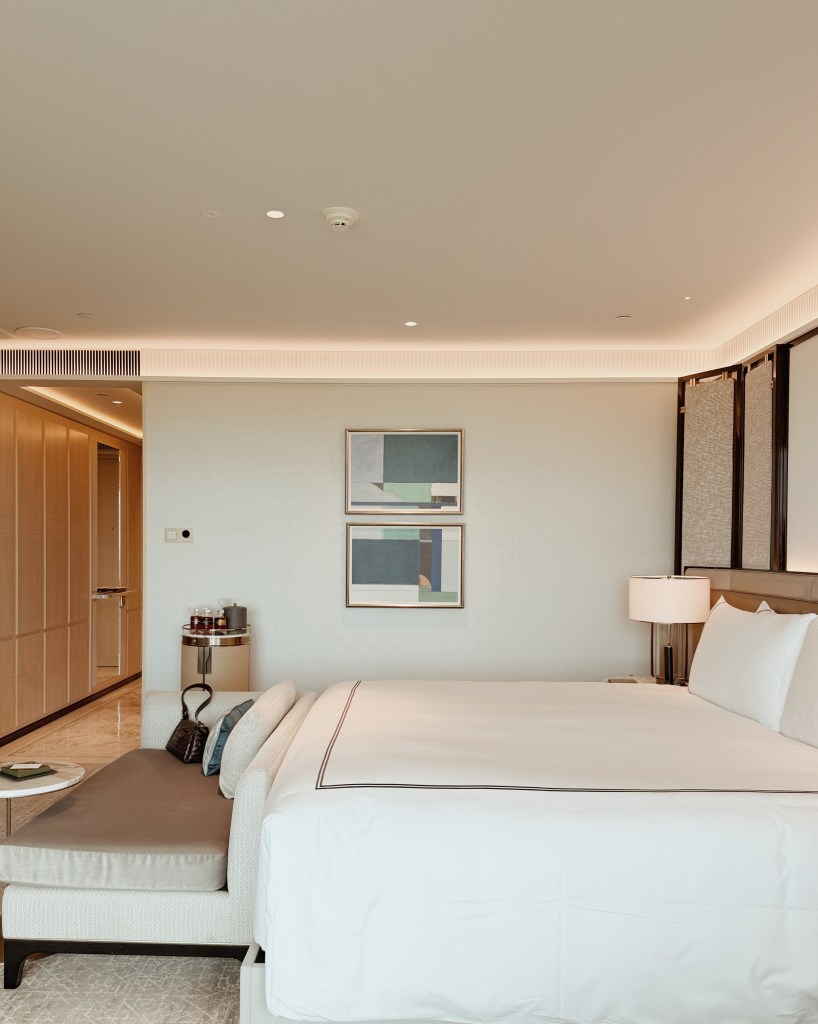 A modern hotel bedroom featuring a large bed with white linens, a stylish seating area, and minimalist decor. Two abstract paintings are displayed on the wall, and there is a bedside lamp and a small table with items on it.