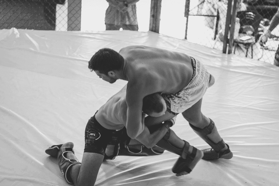 An MMA fighter attempting a takedown on an opponent during training inside a fenced cage.