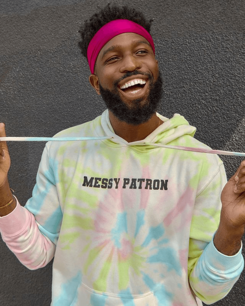 A smiling man with a beard wearing a pastel tie-dye hoodie with "Messy Patron" text and a pink headband.