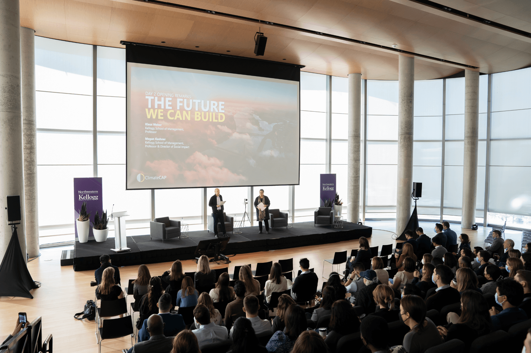 A conference room filled with an audience facing a large screen displaying a presentation. Bright windows in the background.